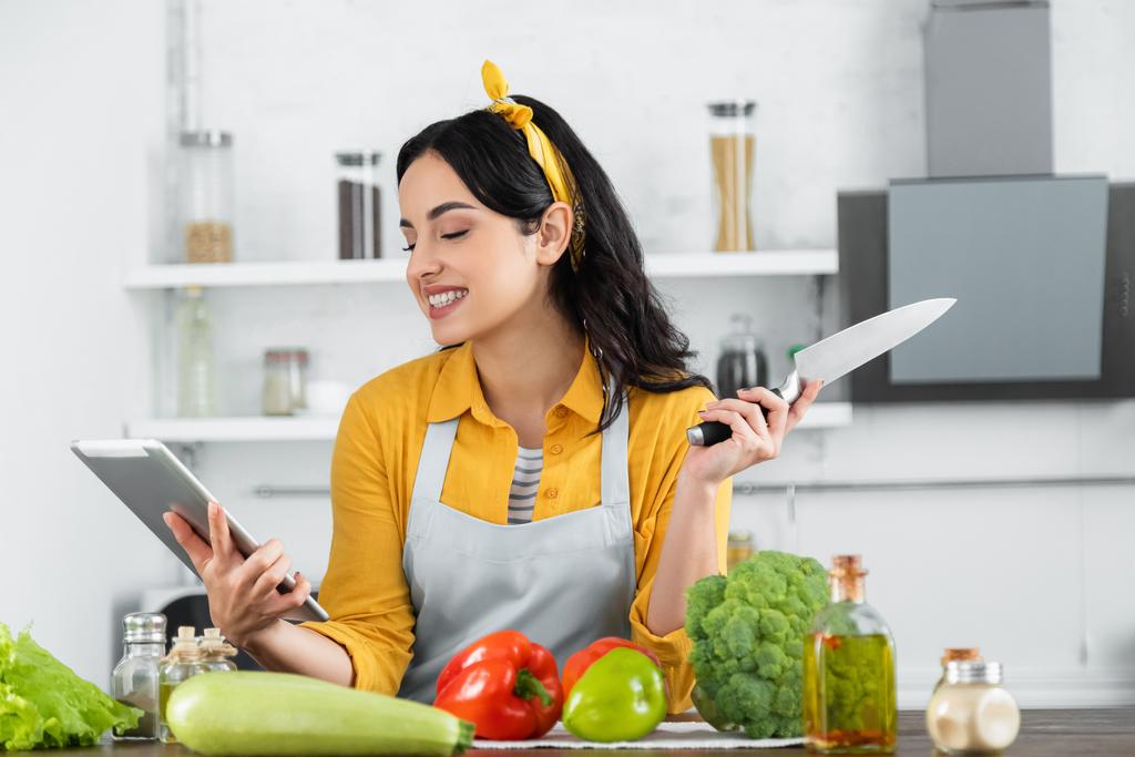 heureuse jeune femme regardant tablette numérique tout en regardant la recette près des légumes sur la table  - Photo, image