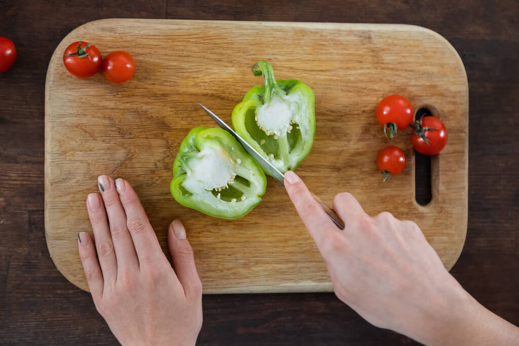 vista superior de la mujer cortando pimiento verde cerca de tomates cherry maduros sobre tabla de cortar de madera  - Foto, Imagen