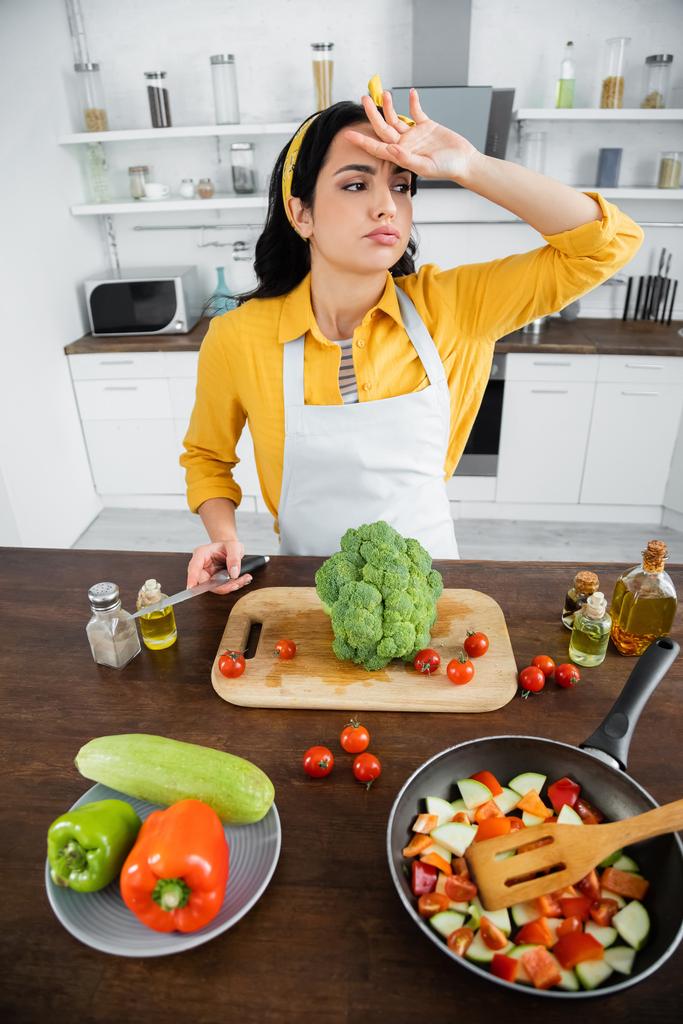 уставшая молодая женщина в фартуке вытирает пот, держа нож рядом с овощами и сковородку на кухне  - Фото, изображение