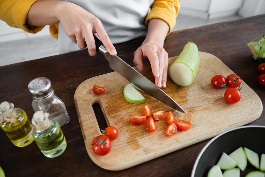 vista parcial de la mujer cortando calabacín cerca de los tomates cherry en rodajas en la tabla de cortar  - Foto, Imagen