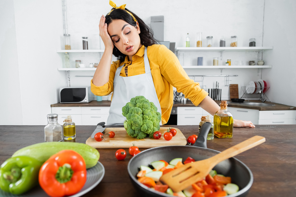 уставшая молодая женщина в фартуке смотрит на овощи возле сковороды на кухне  - Фото, изображение