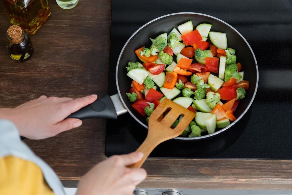 высокий угол обзора молодой женщины, смешивающей нарезанные овощи на сковороде с лопаткой  - Фото, изображение