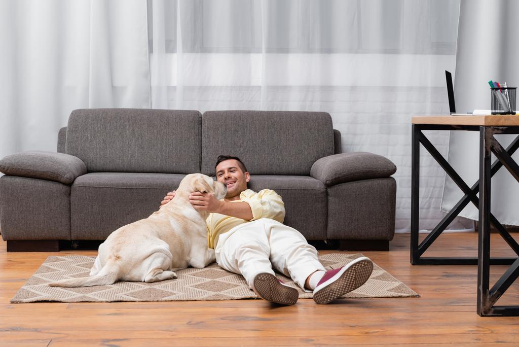 young man having fun with labrador dog while lying on floor near sofa - Photo, Image