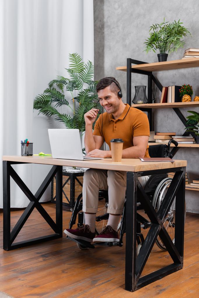 alegre, hombre discapacitado en auriculares trabajando en la oficina en casa cerca de la computadora portátil - Foto, Imagen