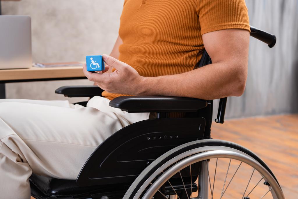 καλλιεργημένη άποψη του άνδρα σε αναπηρικό αμαξίδιο κρατώντας κύβο με το σήμα αναπηρίας - Φωτογραφία, εικόνα