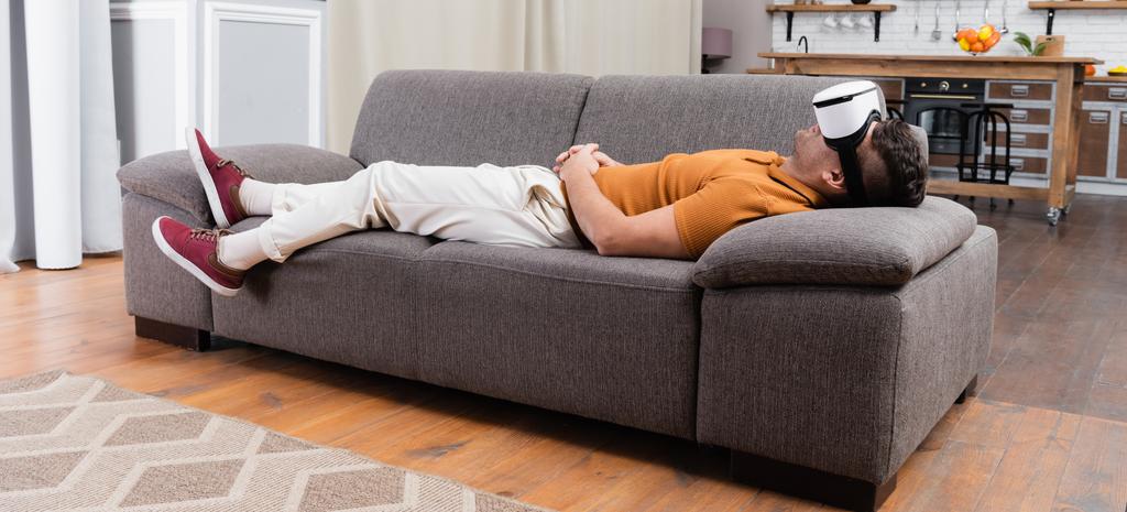 jeune homme en vêtements décontractés couché dans le casque vr sur le canapé à la maison, bannière - Photo, image