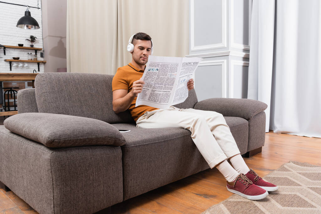 hombre en auriculares escuchando música y leyendo periódicos en el sofá en casa - Foto, Imagen