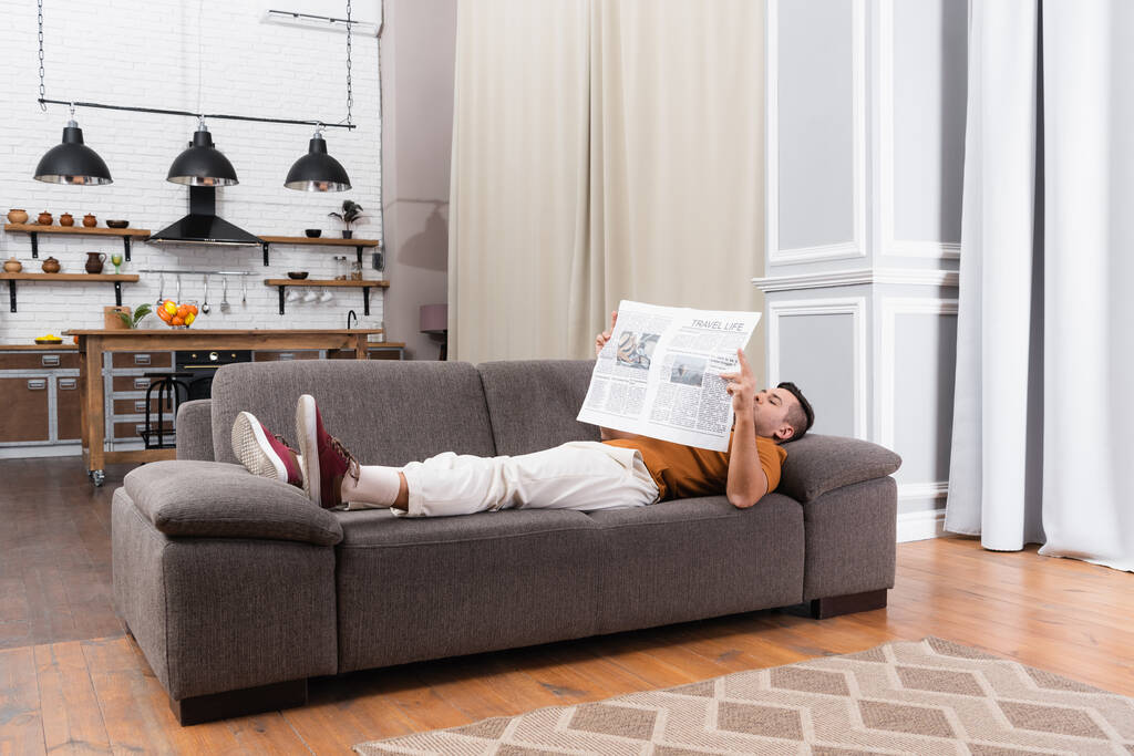 joven leyendo el periódico mientras está acostado en un sofá en un apartamento moderno - Foto, imagen