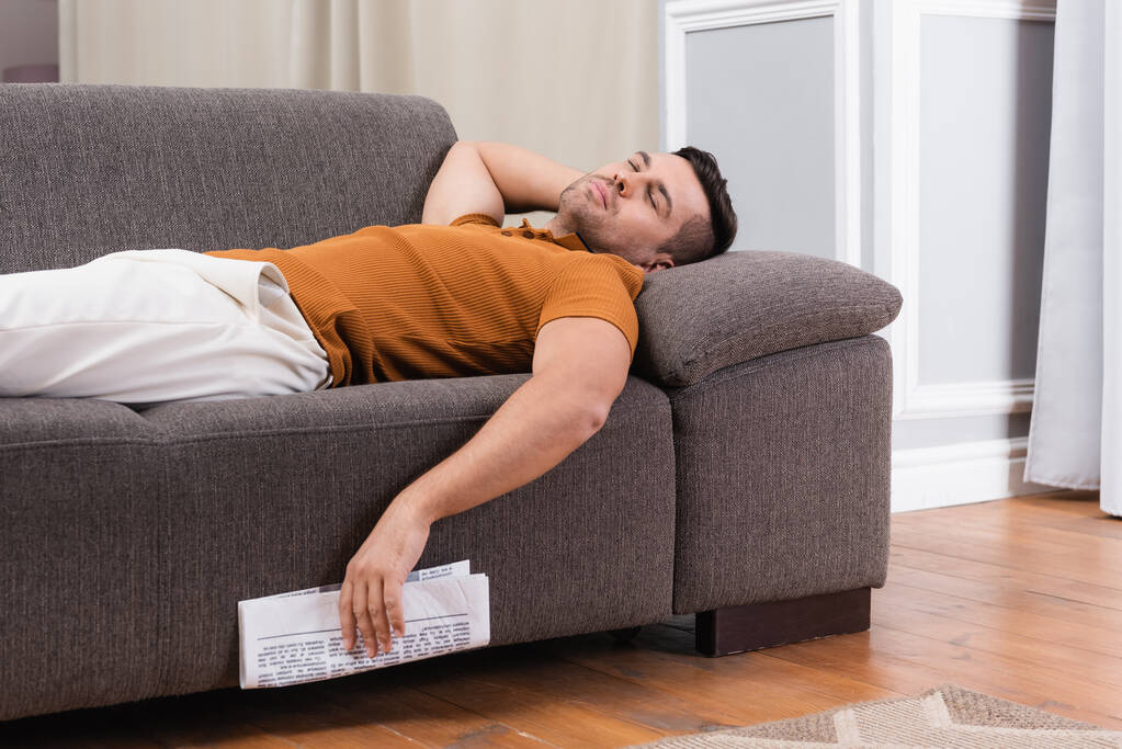 Молодой человек спит на диване, держа газету - Фото, изображение