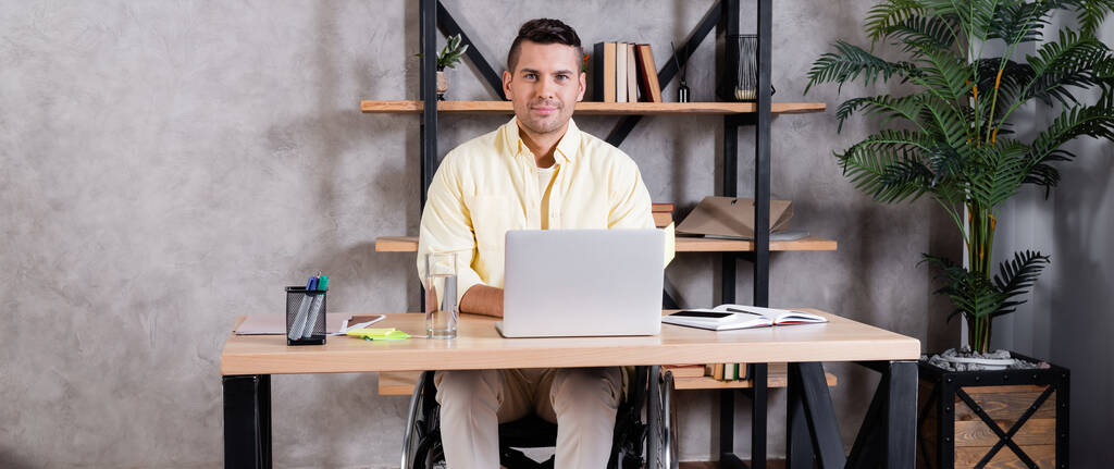 hombre discapacitado sonriendo a la cámara mientras trabaja cerca de la computadora portátil en casa, pancarta - Foto, Imagen