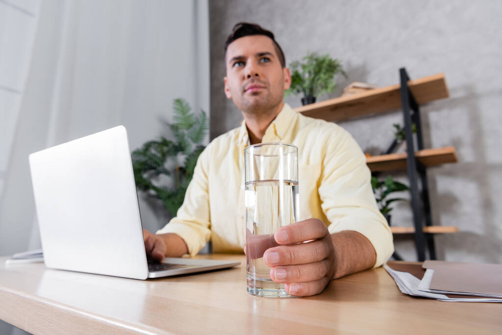 foyer sélectif de verre avec de l'eau en main de l'homme travaillant près d'un ordinateur portable sur fond flou - Photo, image