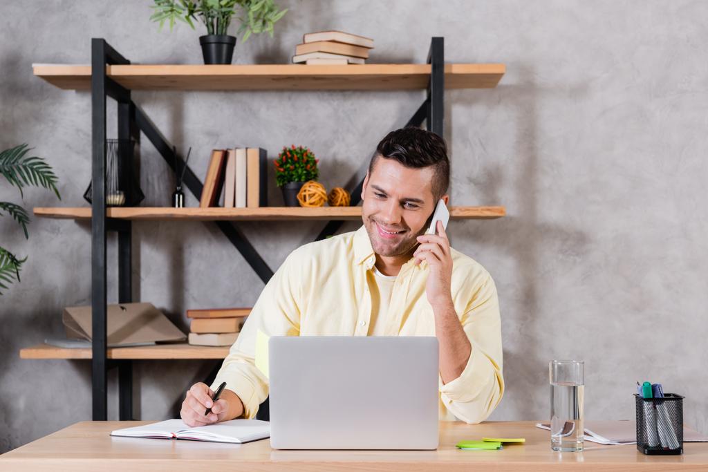 uśmiechnięty mężczyzna rozmawiający przez telefon komórkowy podczas pracy w pobliżu laptopa w domu - Zdjęcie, obraz