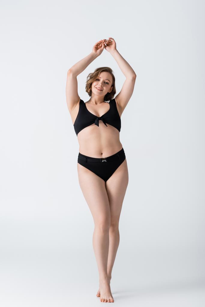 full length of overweight and barefoot young woman smiling while posing with raised hands on white  - Photo, Image