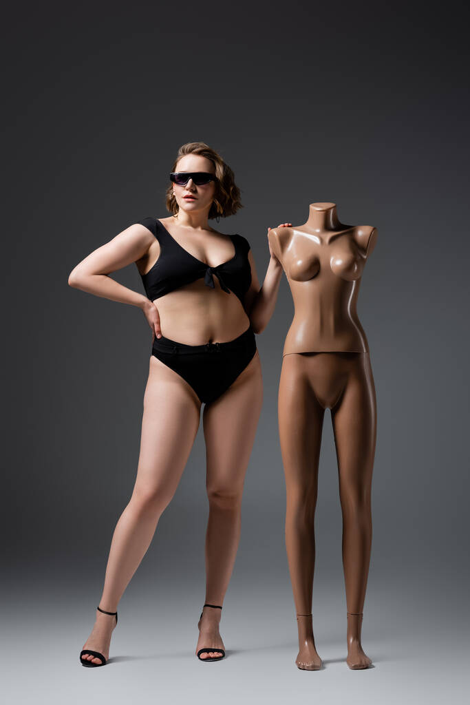 longitud completa de la mujer joven con sobrepeso en traje de baño y gafas de sol posando con maniquí de plástico en gris - Foto, Imagen