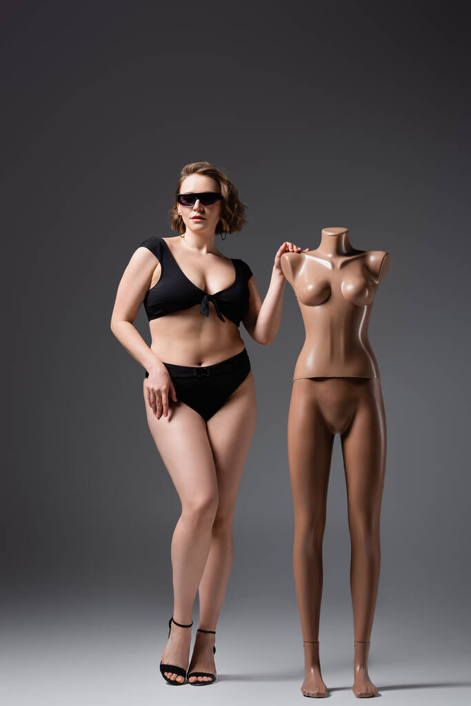 πλήρες μήκος της υπέρβαρη νεαρή γυναίκα σε μαγιό και γυαλιά ηλίου ποζάρουν κοντά σε πλαστική κούκλα σε γκρι - Φωτογραφία, εικόνα