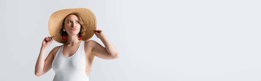 jeune femme en surpoids en boucles d'oreilles et maillot de bain ajustant chapeau de paille et lèvres boudantes isolées sur blanc, bannière - Photo, image