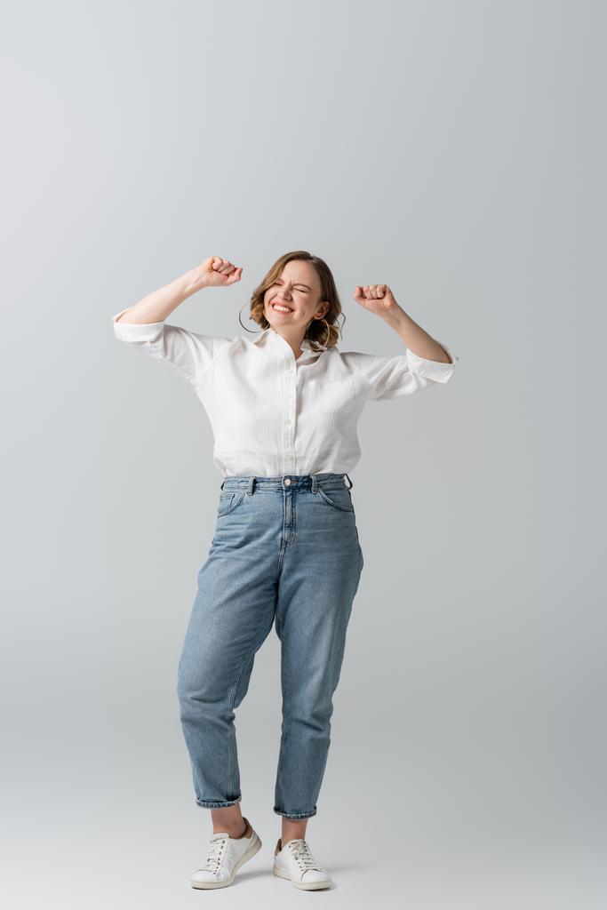 plná délka potěšené nadváhy ženy v džínách slaví na šedé  - Fotografie, Obrázek