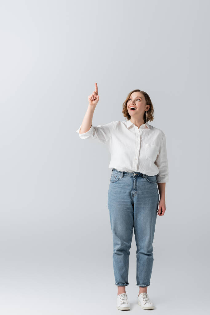 longitud completa de la mujer feliz con sobrepeso en jeans señalando con el dedo en gris  - Foto, Imagen