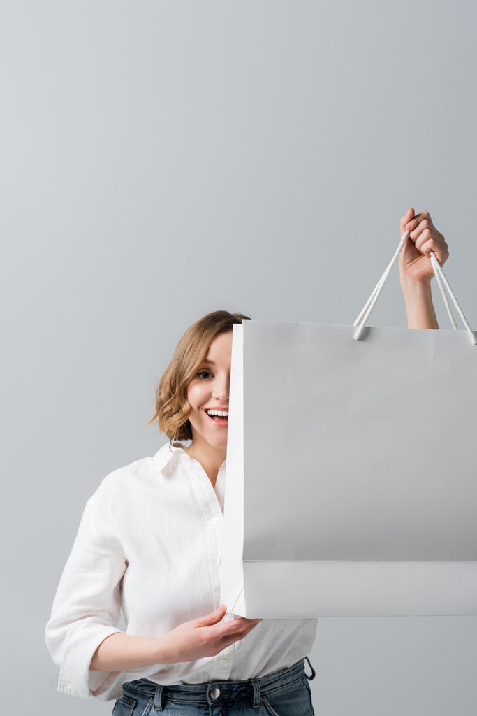 ευτυχισμένη υπέρβαρη γυναίκα σε λευκό πουκάμισο κρατώντας τσάντα ψώνια σε γκρι  - Φωτογραφία, εικόνα