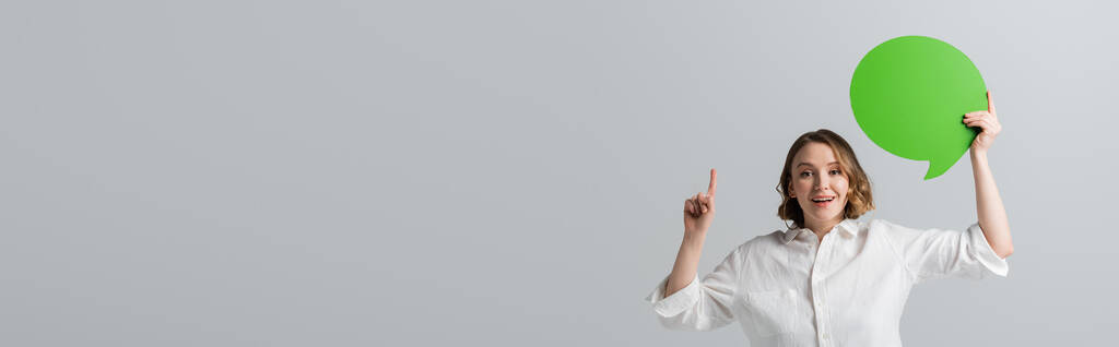Beyaz tişörtlü, yeşil konuşma baloncuğu tutan ve gri pankartta izole edilmiş parmağıyla işaret eden mutlu şişman kadın. - Fotoğraf, Görsel