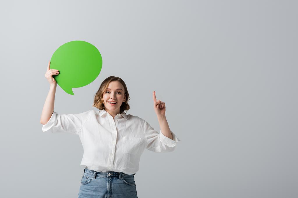 gelukkig overgewicht vrouw in wit shirt met groene spraak zeepbel en wijzen met de vinger geïsoleerd op grijs  - Foto, afbeelding