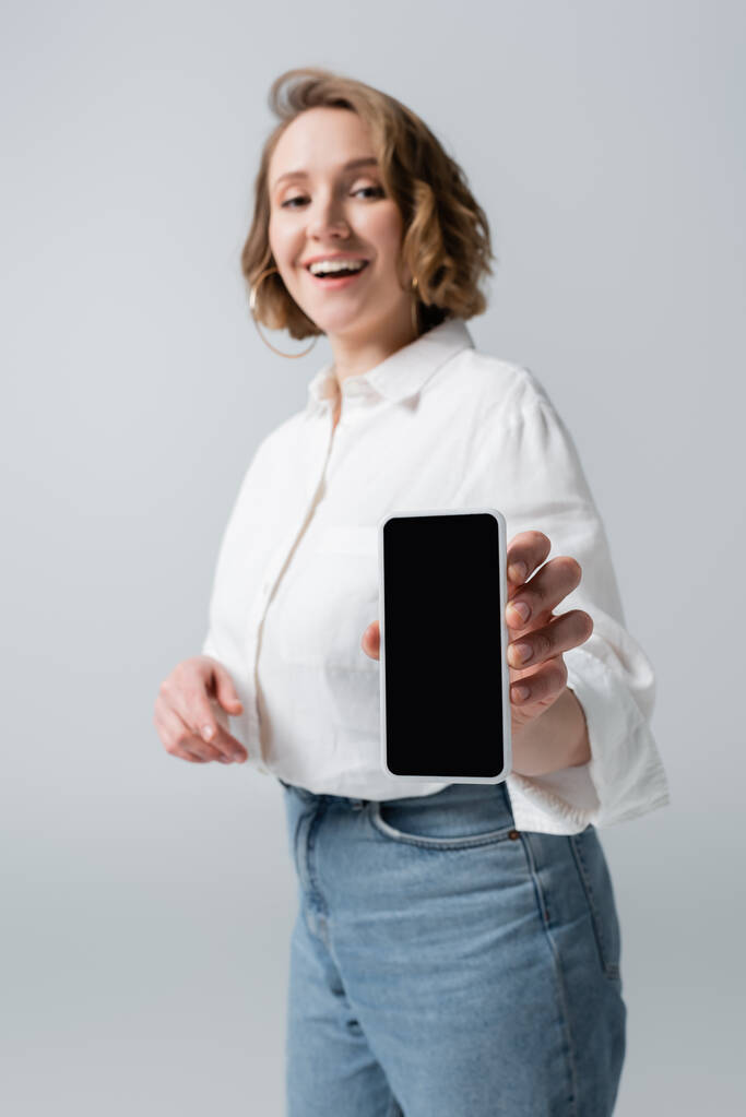 θολή υπέρβαρη γυναίκα κρατώντας smartphone με κενή οθόνη και χαμογελώντας απομονωμένη σε γκρι - Φωτογραφία, εικόνα