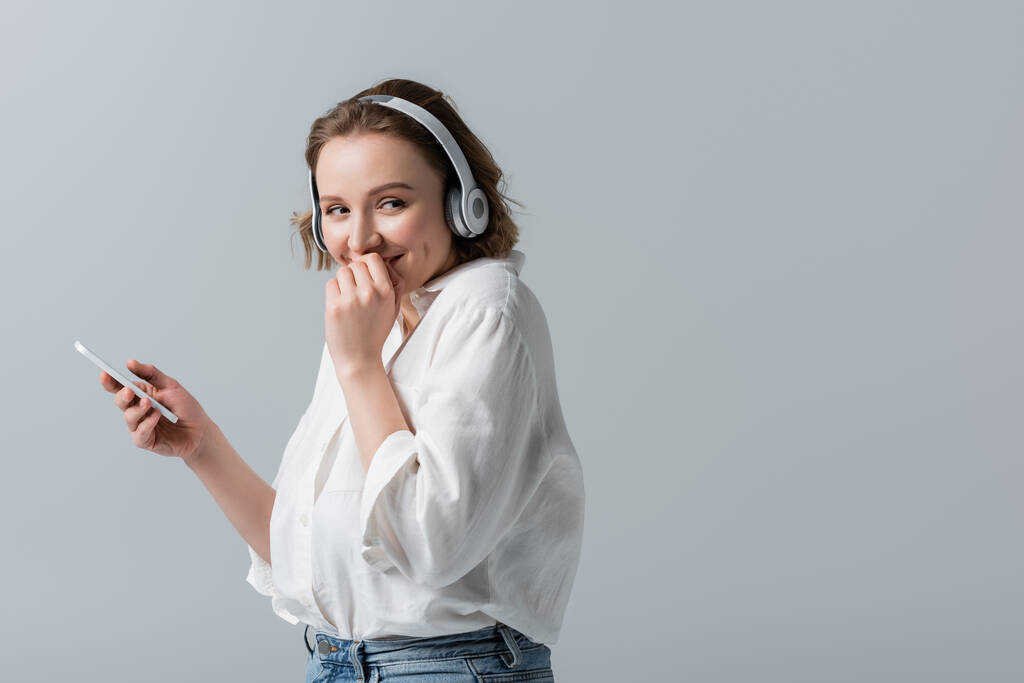счастливая плюс размер женщина в беспроводных наушниках прослушивания музыки и проведения смартфона изолированы на серый - Фото, изображение