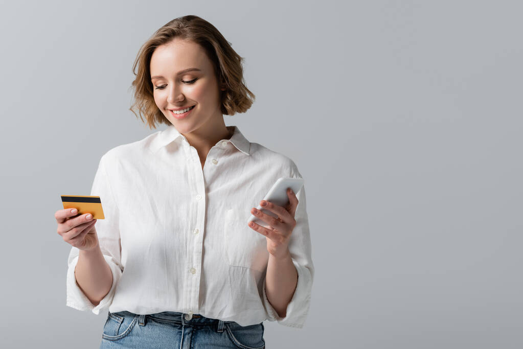 alegre más tamaño mujer celebración de tarjeta de crédito y teléfono inteligente aislado en gris - Foto, imagen