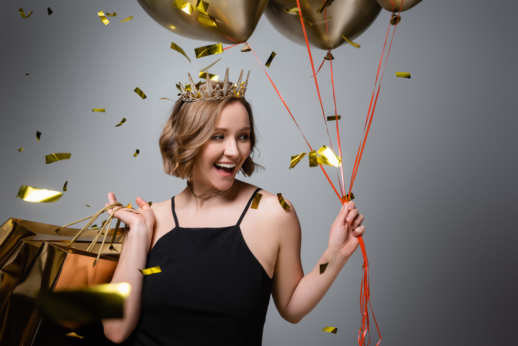 happy plus size woman in slip dress and crown holding balloons and shopping bags near confetti on grey  - Photo, Image