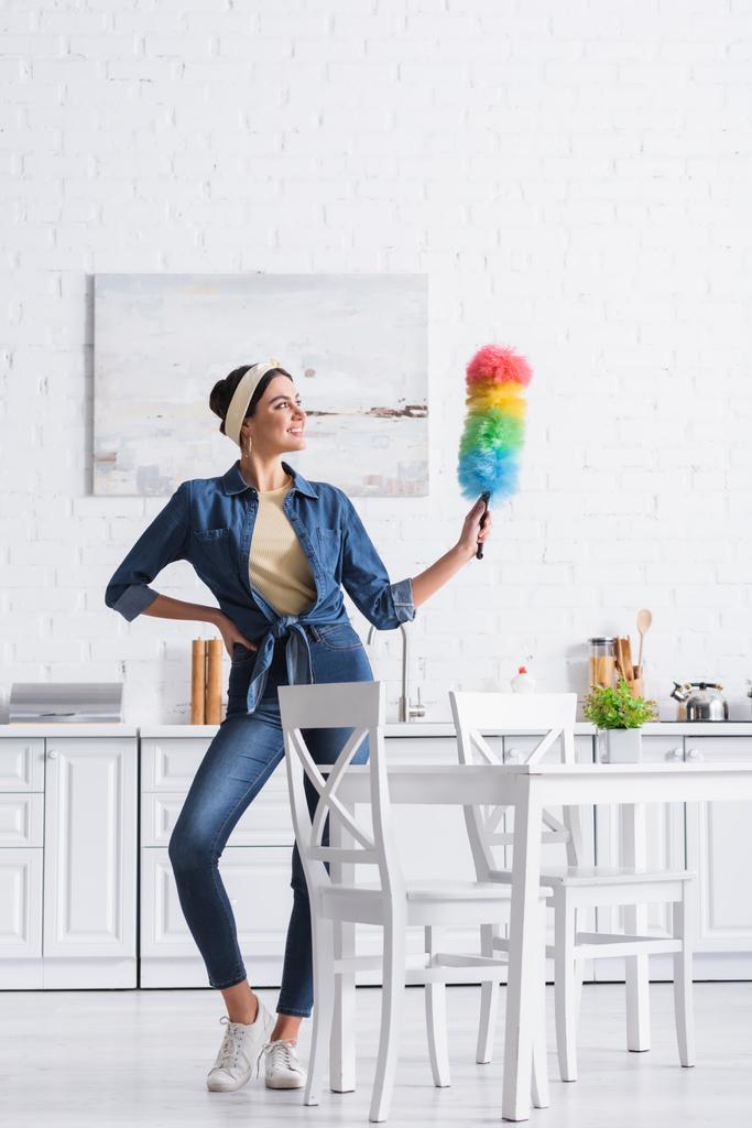 Mujer sonriente en diadema sosteniendo cepillo de polvo en la cocina  - Foto, imagen
