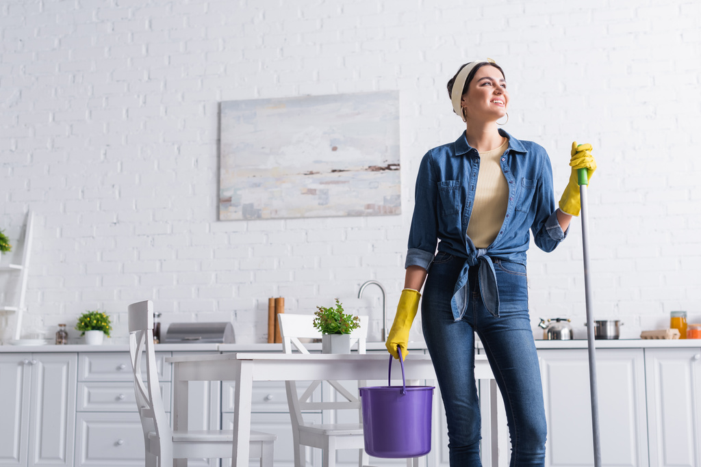 Mulher alegre em luvas de borracha segurando esfregona e balde na cozinha  - Foto, Imagem