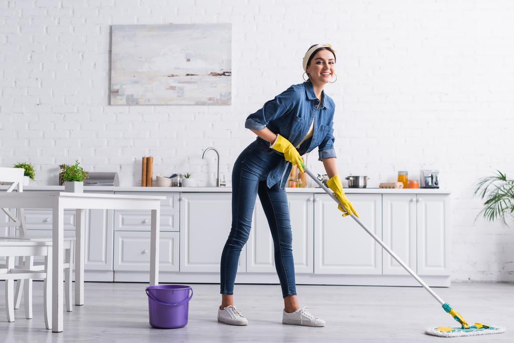 Young woman smiling while washing floor with mop in kitchen  - Photo, Image