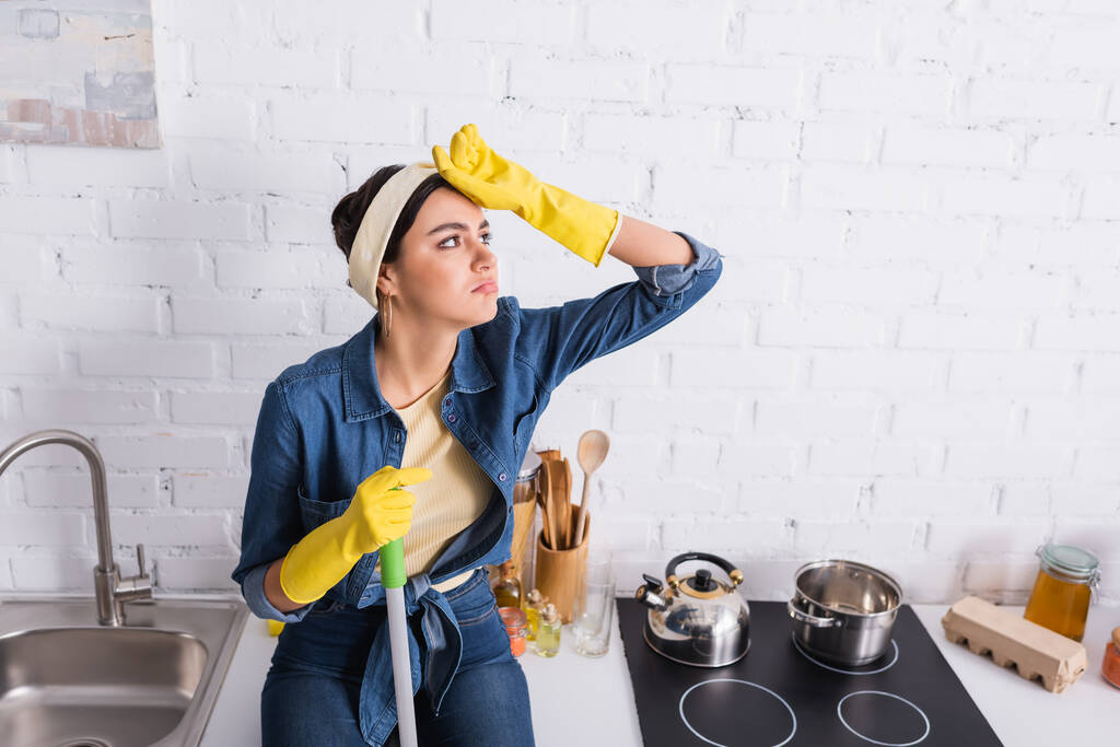 Agotado ama de casa en guantes de goma sosteniendo trapeador en encimera de cocina  - Foto, Imagen