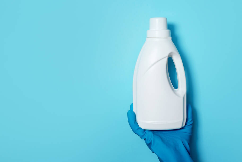 Hand in glove holding white plastic bottle of cleaning product, household chemicals. Copy space. Cleaning service concept. Household chemical cleaning products, brushes and supplies. Detergent bottle - Photo, Image