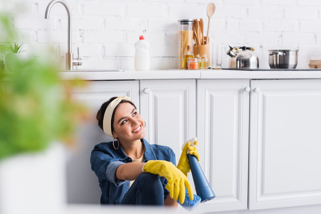 Cheerful woman in rubber gloves holding bottle of detergent and sponge in kitchen  - Photo, Image