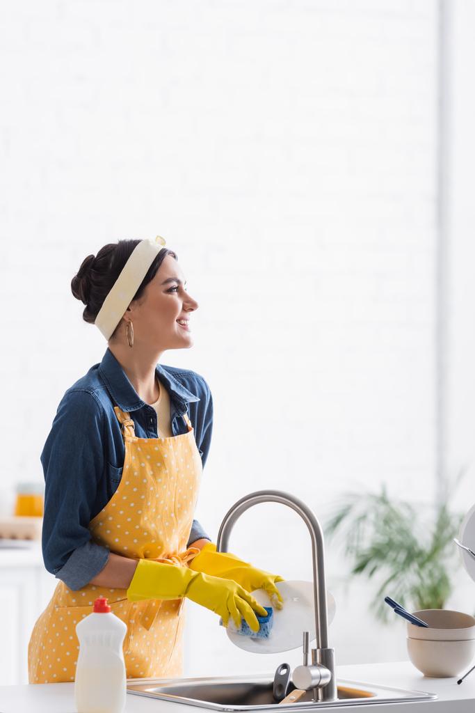 Vrolijke vrouw in schort en rubberen handschoenen afwasbord in keuken  - Foto, afbeelding