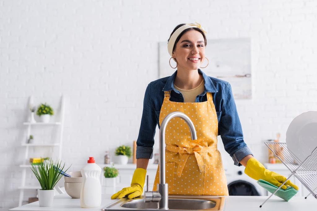 Uśmiechnięta kobieta trzyma szmatę przy talerzach i tonie w kuchni  - Zdjęcie, obraz