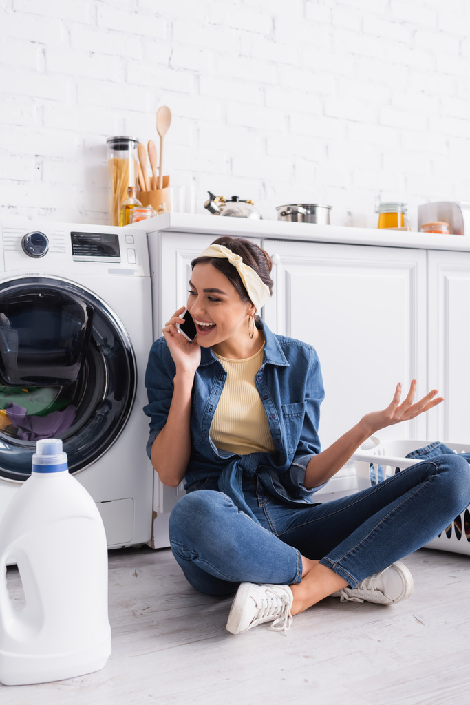 Positiivinen kotiäiti puhuu älypuhelimella pesuaineen ja pesukoneen lähellä  - Valokuva, kuva