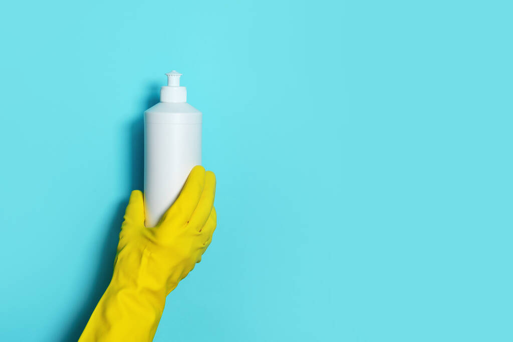 Рука в рукавичці тримає білу пластикову пляшку для очищення продукту, побутові хімічні речовини. Копіювати простір. Концепція прибирання. Побутові хімічні засоби для чищення, щітки та приладдя. Пляшка для миючих засобів
 - Фото, зображення