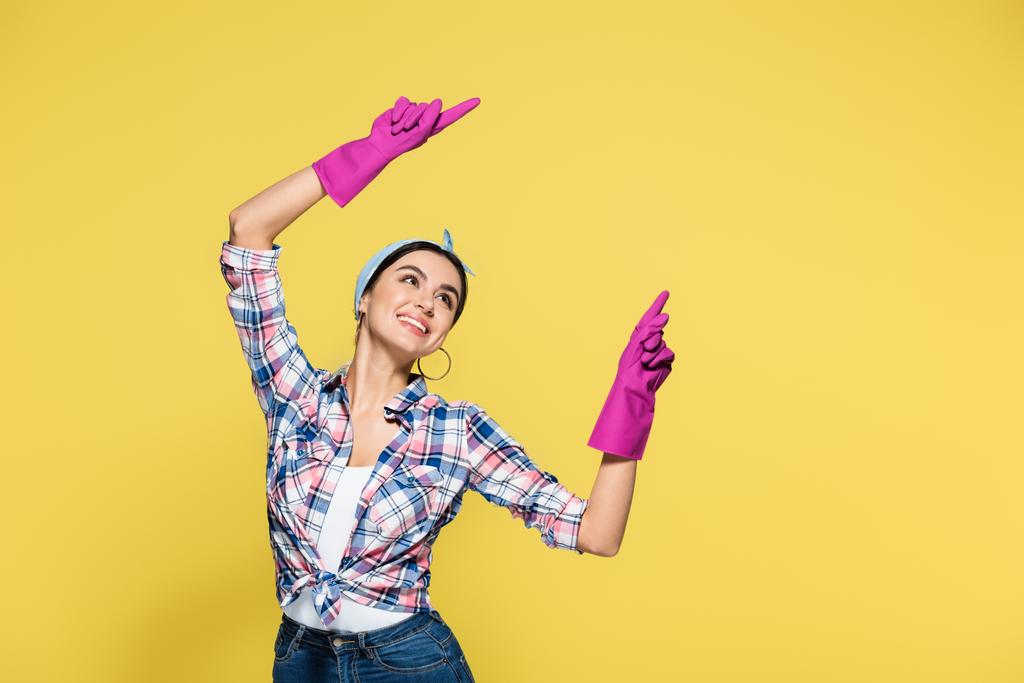 Χαρούμενη νεαρή νοικοκυρά με λαστιχένια γάντια που δείχνει με τα δάχτυλα απομονωμένα στο κίτρινο  - Φωτογραφία, εικόνα