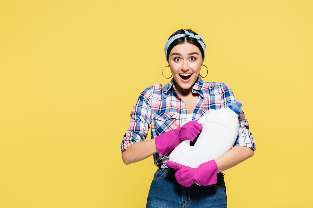 Surprised woman in rubber gloves holding bottle of detergent isolated on yellow  - Photo, Image