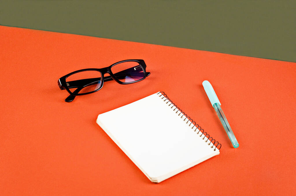 Biały notatnik, okulary i długopis na pomarańczowym i zielonym tle. Minimalistyczna koncepcja z miejscem na tekst. Makieta z elementami dekoracyjnymi - Zdjęcie, obraz