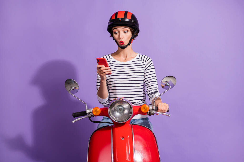 Foto de sorprendida señora loca paseo moto celebrar el aspecto del teléfono celular pantalla usar casco a rayas camisa aislado color violeta fondo - Foto, imagen