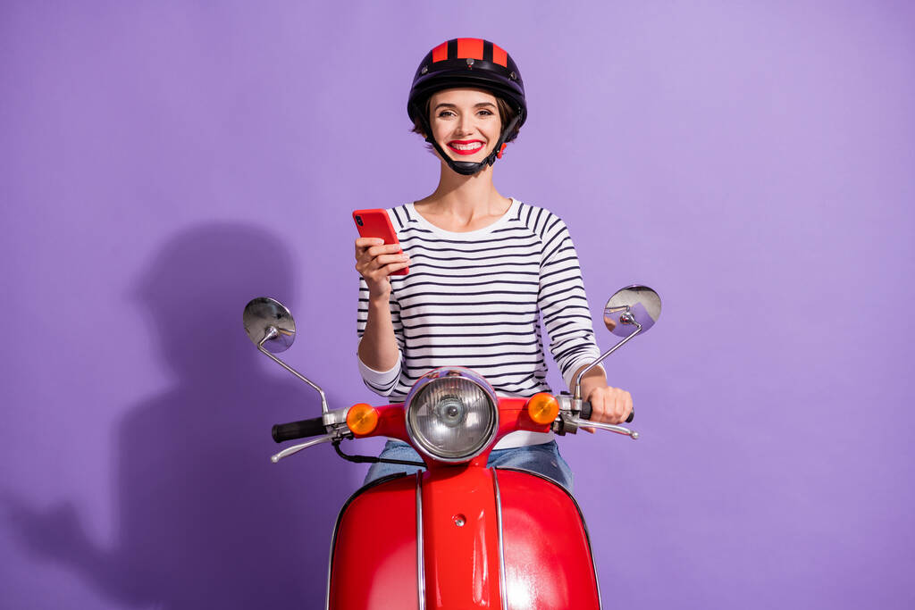 Fénykép a pozitív lány meghajtó moped hold telefon kopás sisak csíkos ing elszigetelt ibolya szín háttér - Fotó, kép