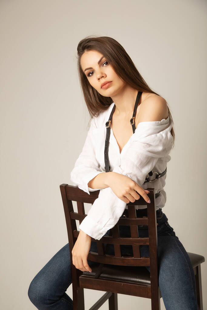 Портрет молодой кавказской брюнетки в белой блузке, джинсах и обвязке, позирующей на стуле и смотрящей в камеру на бежевом фоне - Фото, изображение