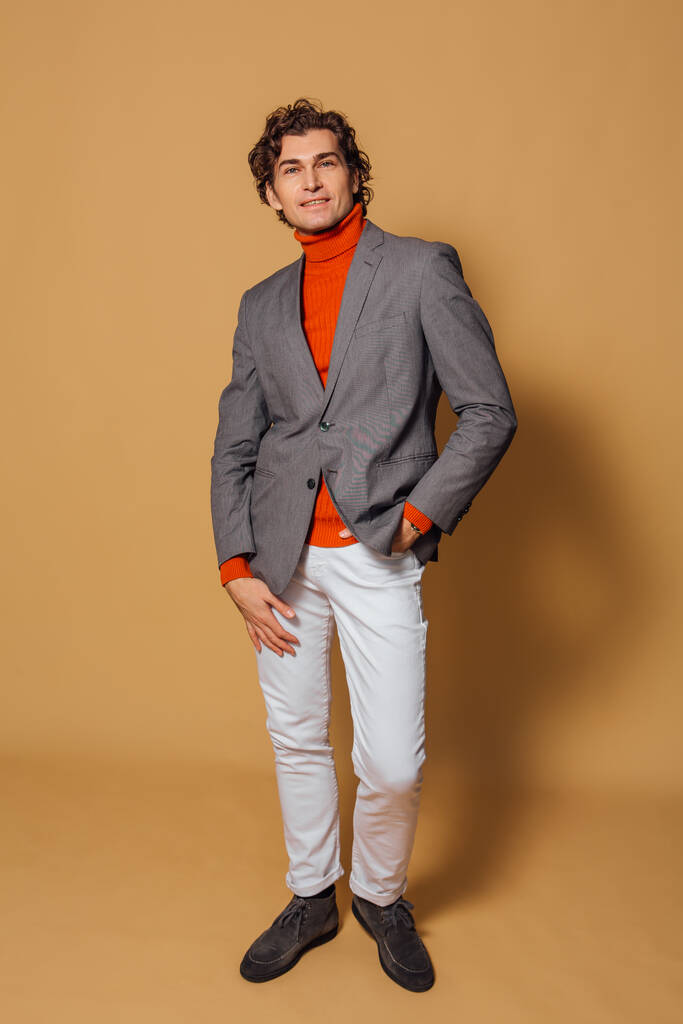 Retrato de moda de un hombre alto y guapo vestido con cuello alto naranja, jeans blancos y chaqueta gris posando sobre el fondo beige - Foto, Imagen