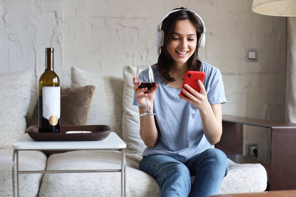 Ładna dziewczyna używająca smartfona na kanapie w domu w salonie. Słuchanie muzyki, picie czerwonego wina, relaks po ciężkim tygodniu w pracy - Zdjęcie, obraz