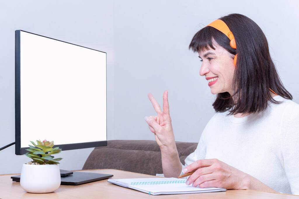 Smiling business woman in headphones looks into a monitor with a blank white screen and shows a victory sign - Photo, Image