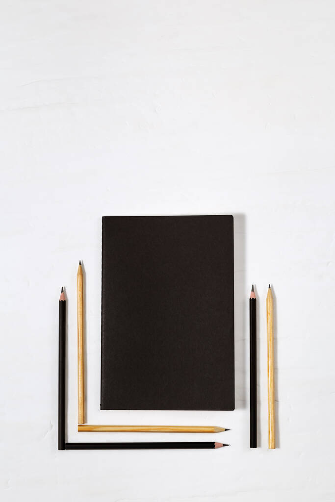 Шесть деревянных карандашей с черным свинцом и закрытой черной книгой. Простые карандаши и макет книги по рабочему пространству стола. Живописные принадлежности с копировальным пространством. Вид сверху. Плоский лежал. - Фото, изображение