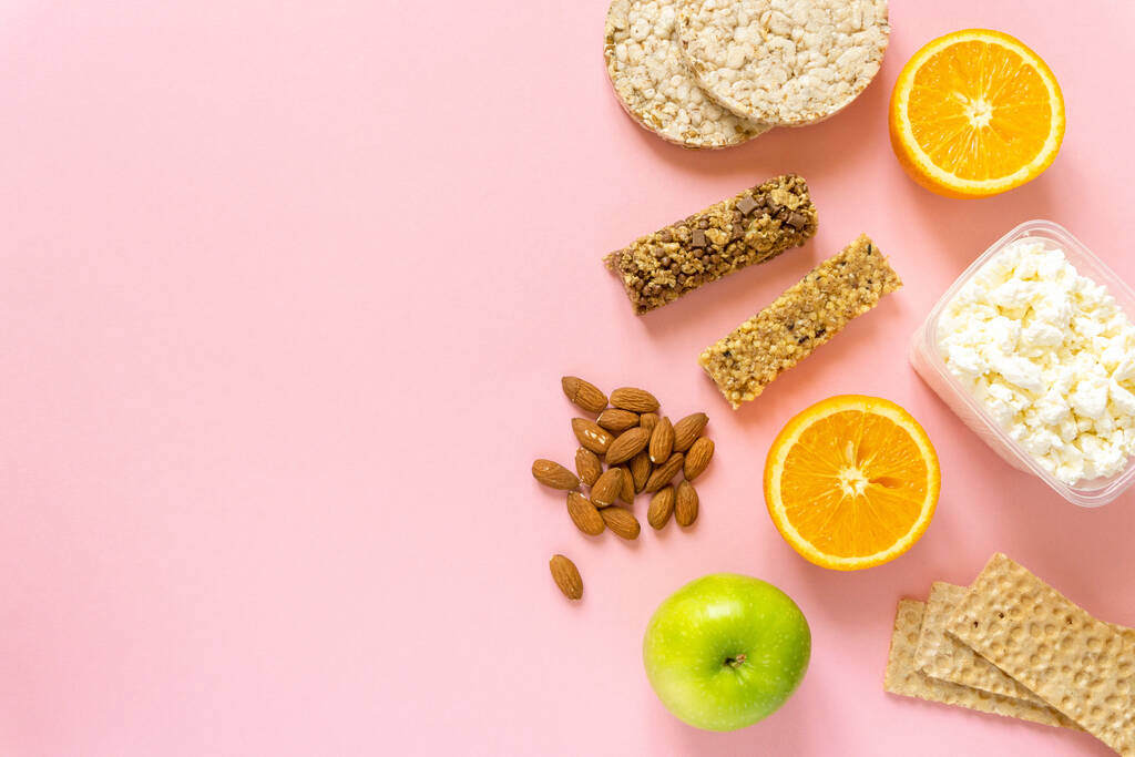 Flat lay of healthy snacks. Fruits, muesli bars, crispbreads, cottage cheese and nuts on pink background, space for text - Photo, Image
